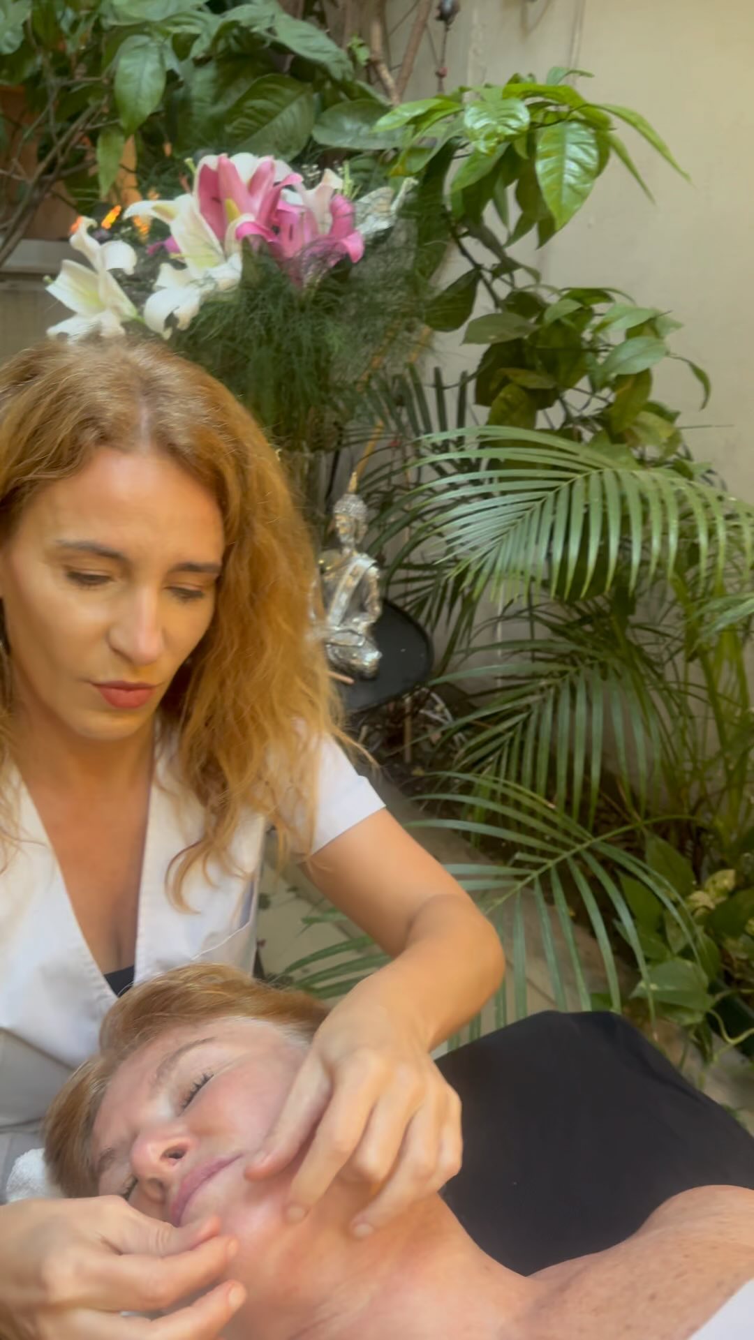 Verónica hace un masaje facial a una clienta recostada, entre plantas y lirios